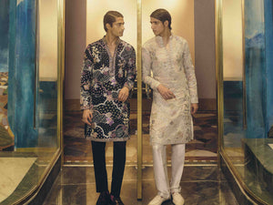 Two men in traditional kurta designs standing in a reflective hallway.